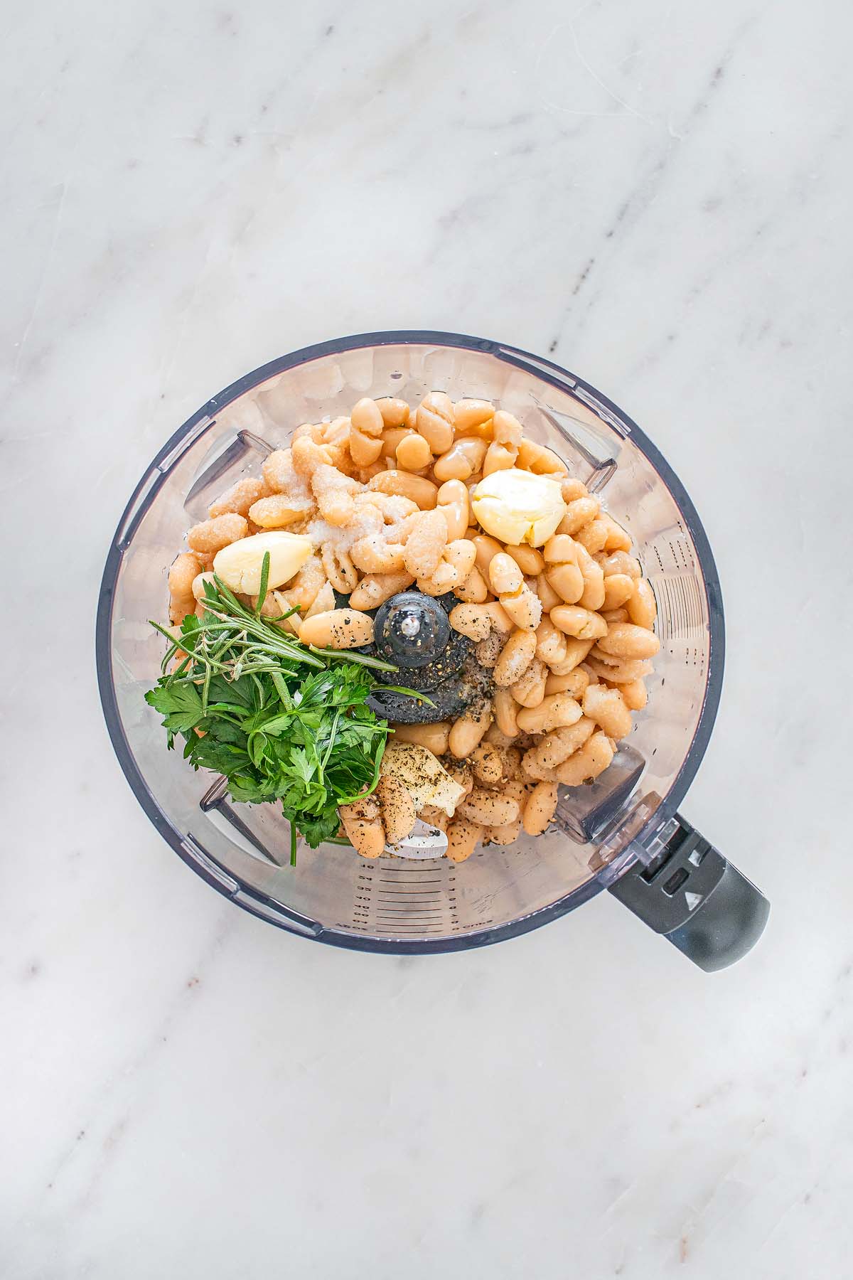 Food processor with white beans, fresh parsley, garlic, black pepper, and olive oil on a marble counter.