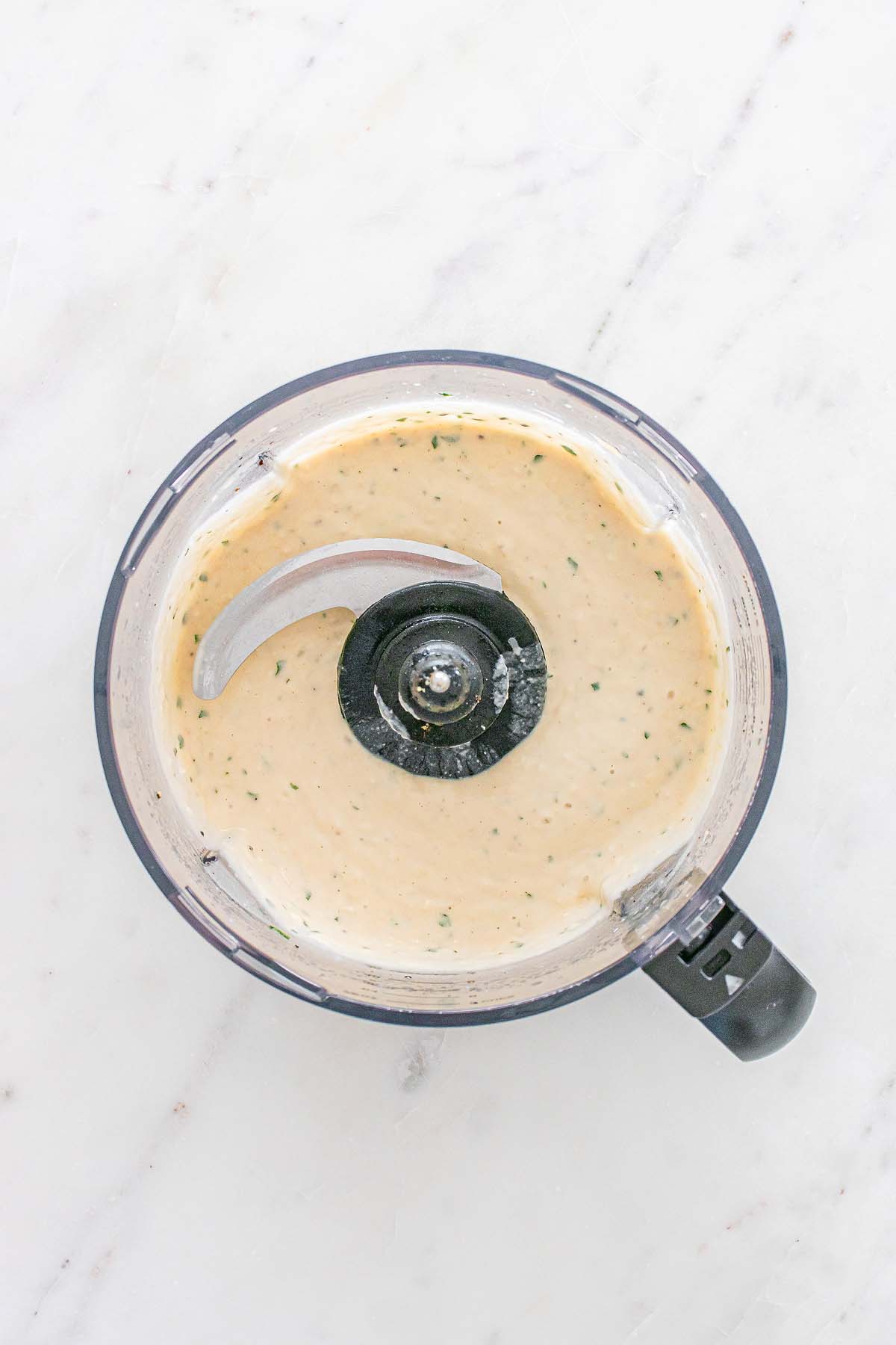 Overhead view of creamy sauce with herbs in a food processor on a white marble surface.