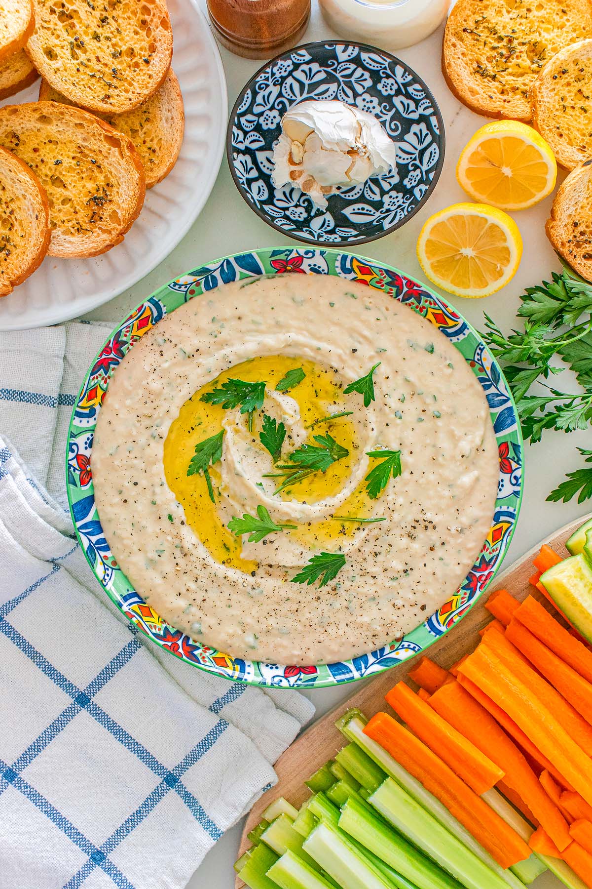 Creamy Italian bean dip garnished with herbs and olive oil, surrounded by toast, lemon, carrot, celery, and parsley.