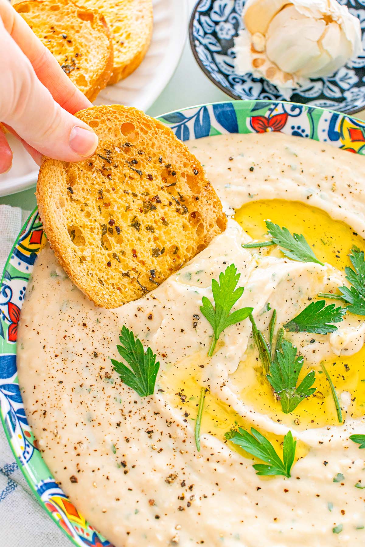 A hand dips toasted bread into creamy cannellini bean topped with olive oil, parsley, and black pepper.
