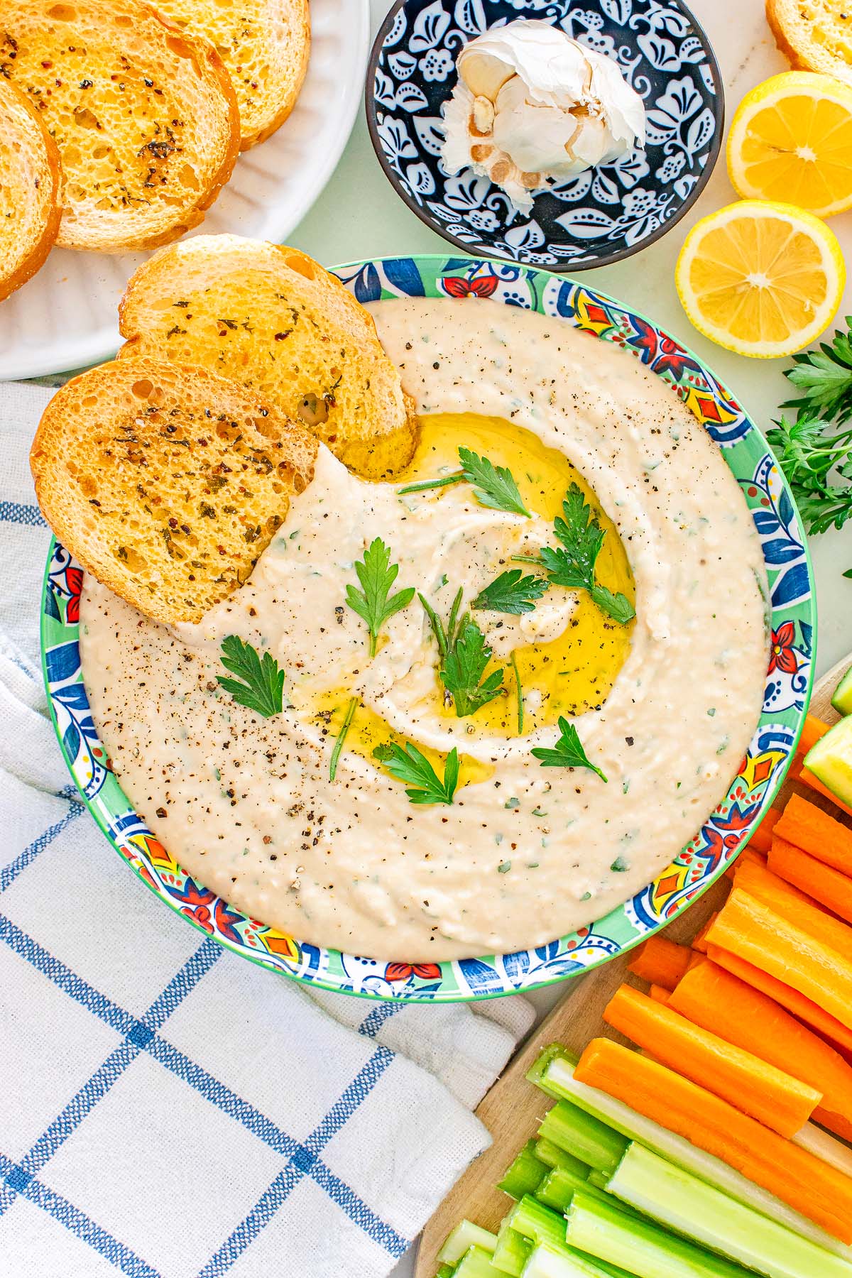 Italian cannellini bean dip in a colorful bowl, topped with parsley and olive oil, served with toast and fresh vegetables.