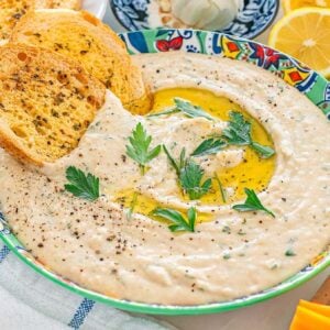 Bowl of creamy white bean dip with herbs and olive oil, surrounded by toast, lemon, and vegetable sticks.