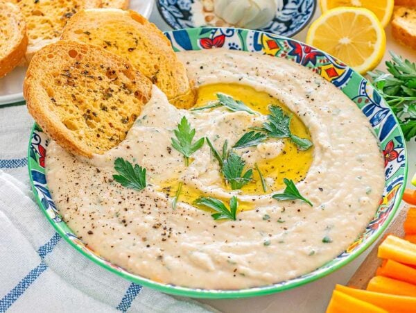 Bowl of creamy white bean dip with herbs and olive oil, surrounded by toast, lemon, and vegetable sticks.