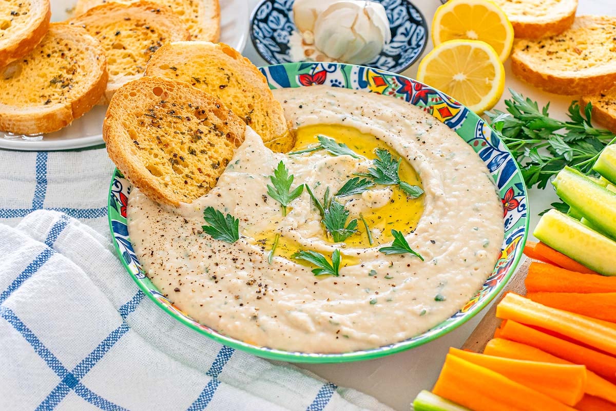 Bowl of creamy white bean dip with herbs and olive oil, surrounded by toast, lemon, and vegetable sticks.
