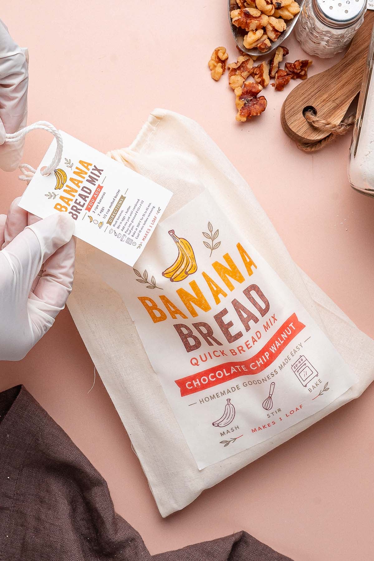 Hands in white gloves holding a cloth bag of Chocolate Chip Walnut Banana Bread mix with an attached recipe tag.