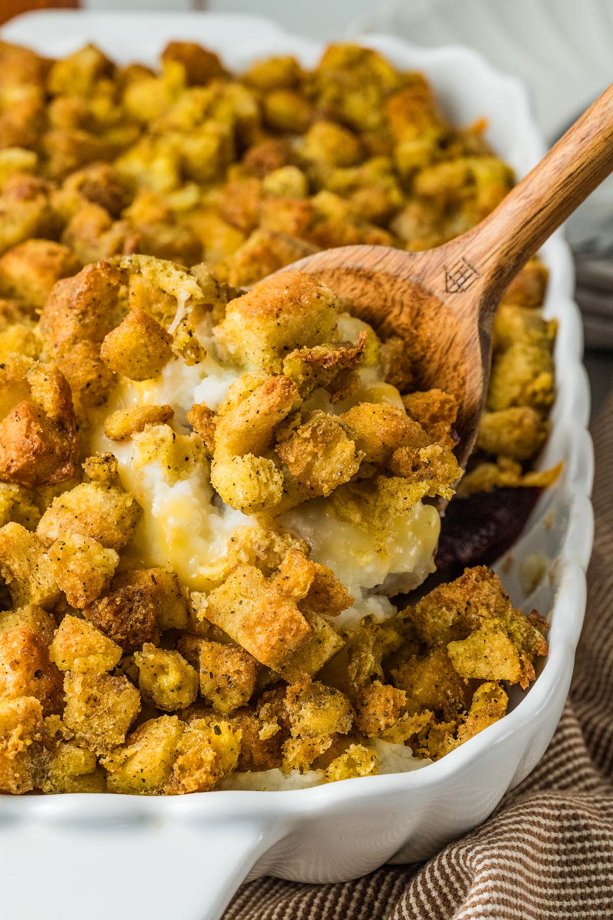 A wooden spoon scooping savory bread stuffing and turkey leftovers out of a white baking dish.