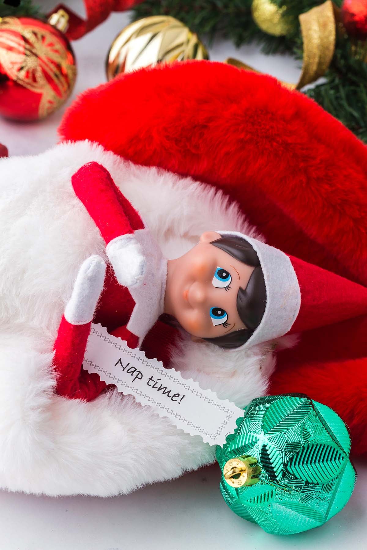 An Elf on the Shelf doll lying on a red and white stocking holding a note that says “Nap time!”.