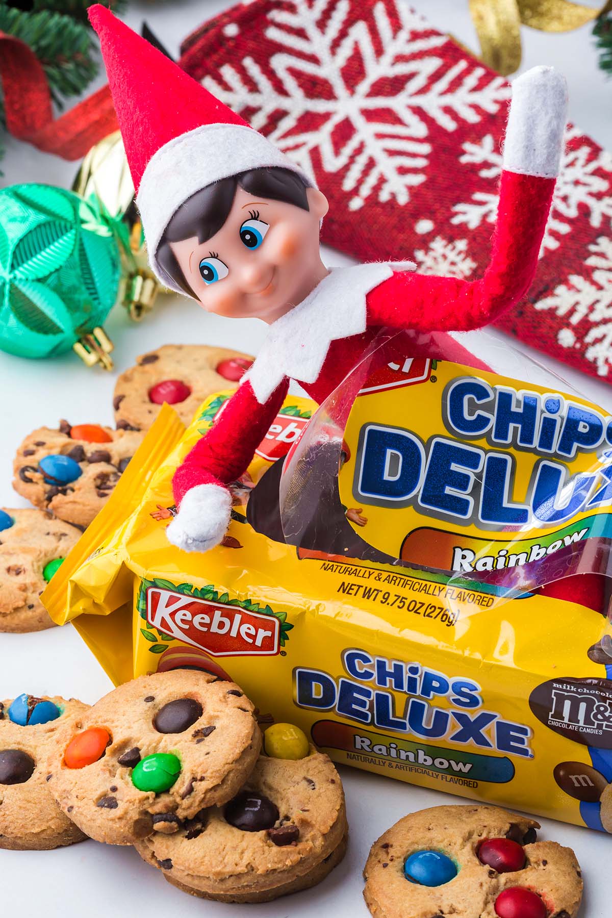 An Elf on the Shelf toy sitting on a yellow package of Keebler Chips Deluxe Rainbow cookies with M&Ms.