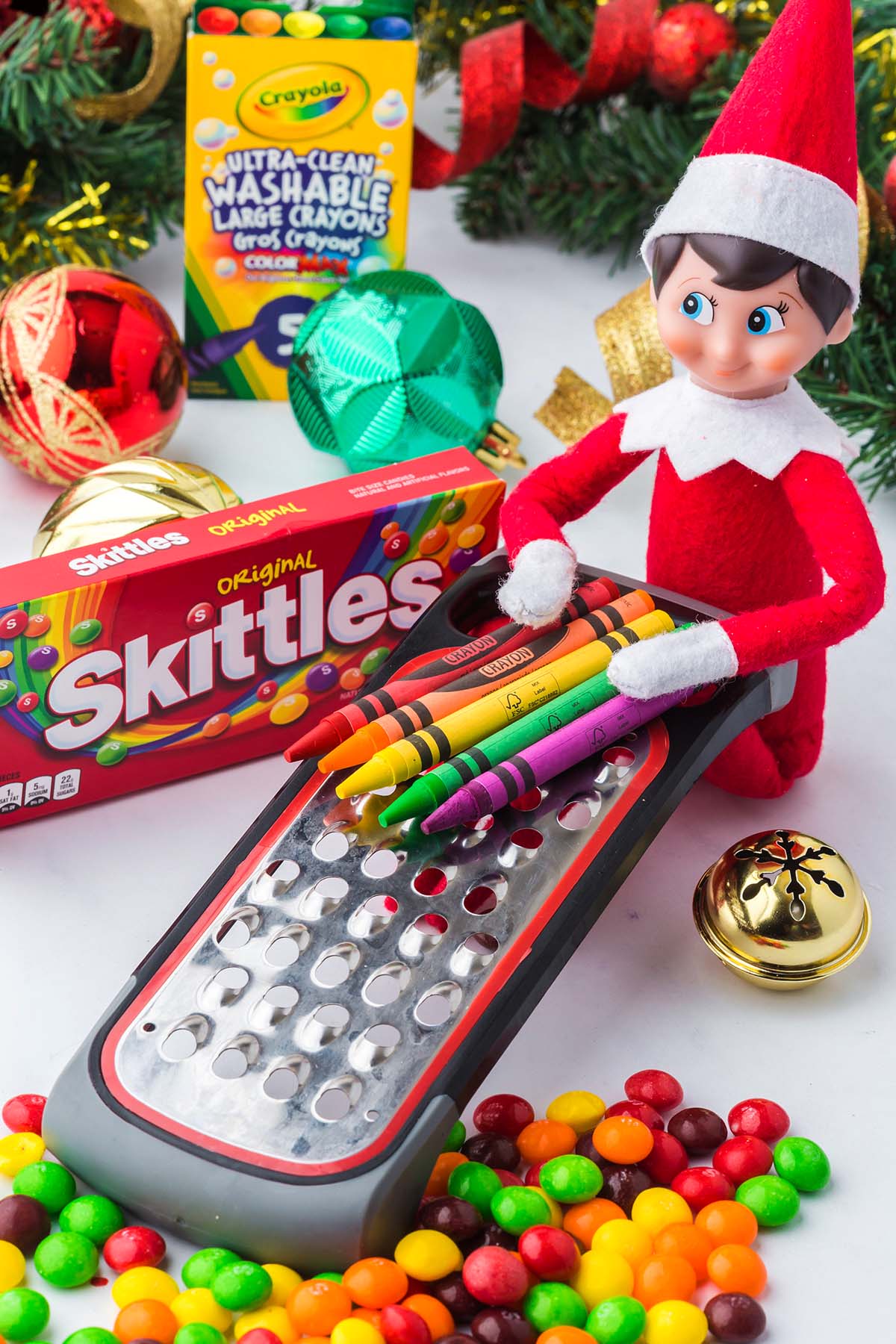 An Elf on the Shelf figurine uses a metal cheese grater to “grate” crayons, with Skittles scattered nearby.