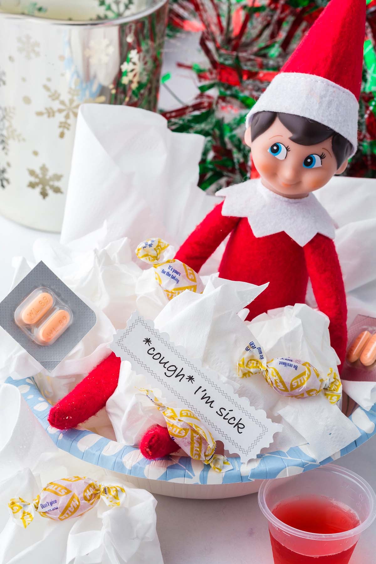 Elf on the Shelf doll sitting in a bowl of tissues with a note that reads “*cough* I’m sick” and cold medication.