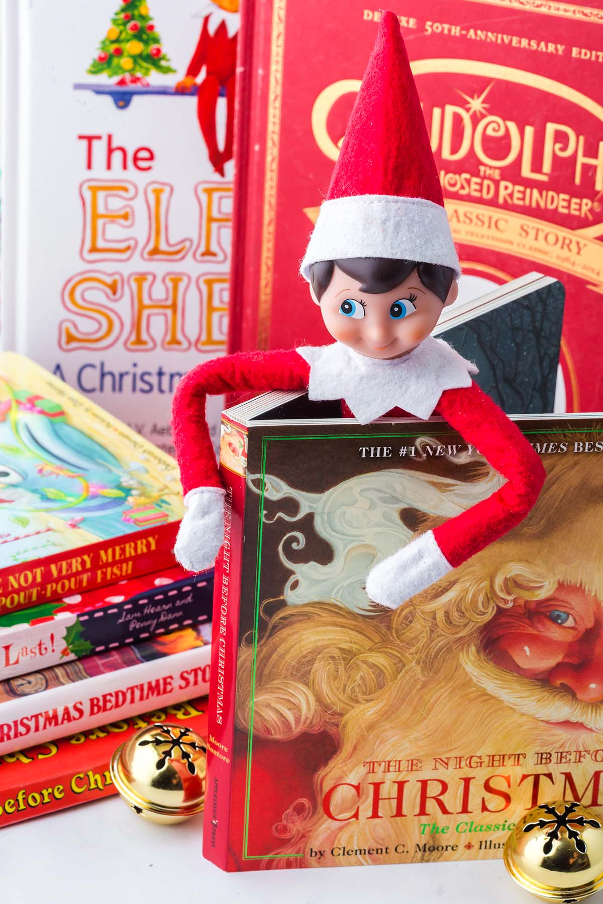 An Elf on the Shelf toy sitting inside an open copy of The Night Before Christmas book, surrounded by other books.