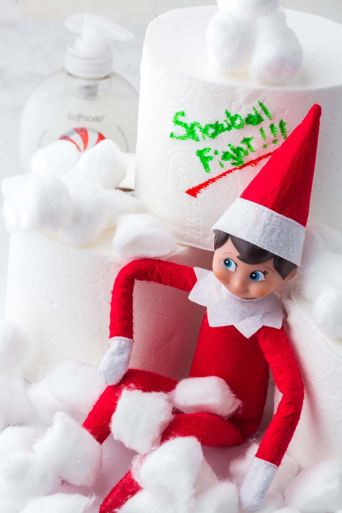 An elf doll sitting in a pile of cotton balls next to toilet paper rolls labeled “Snowball Fight!!!”.