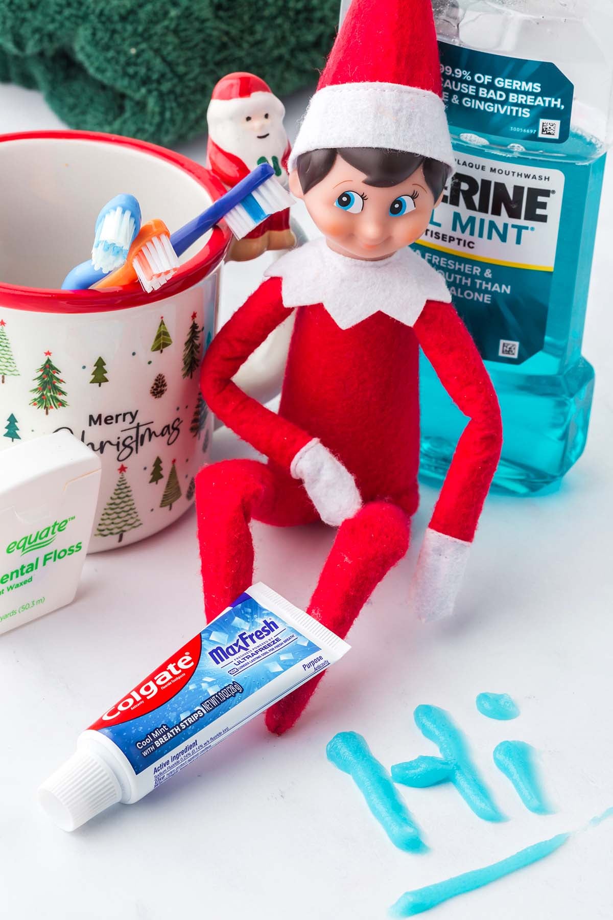 An Elf on the Shelf doll sits among dental products, with the word “hi” spelled out in toothpaste.