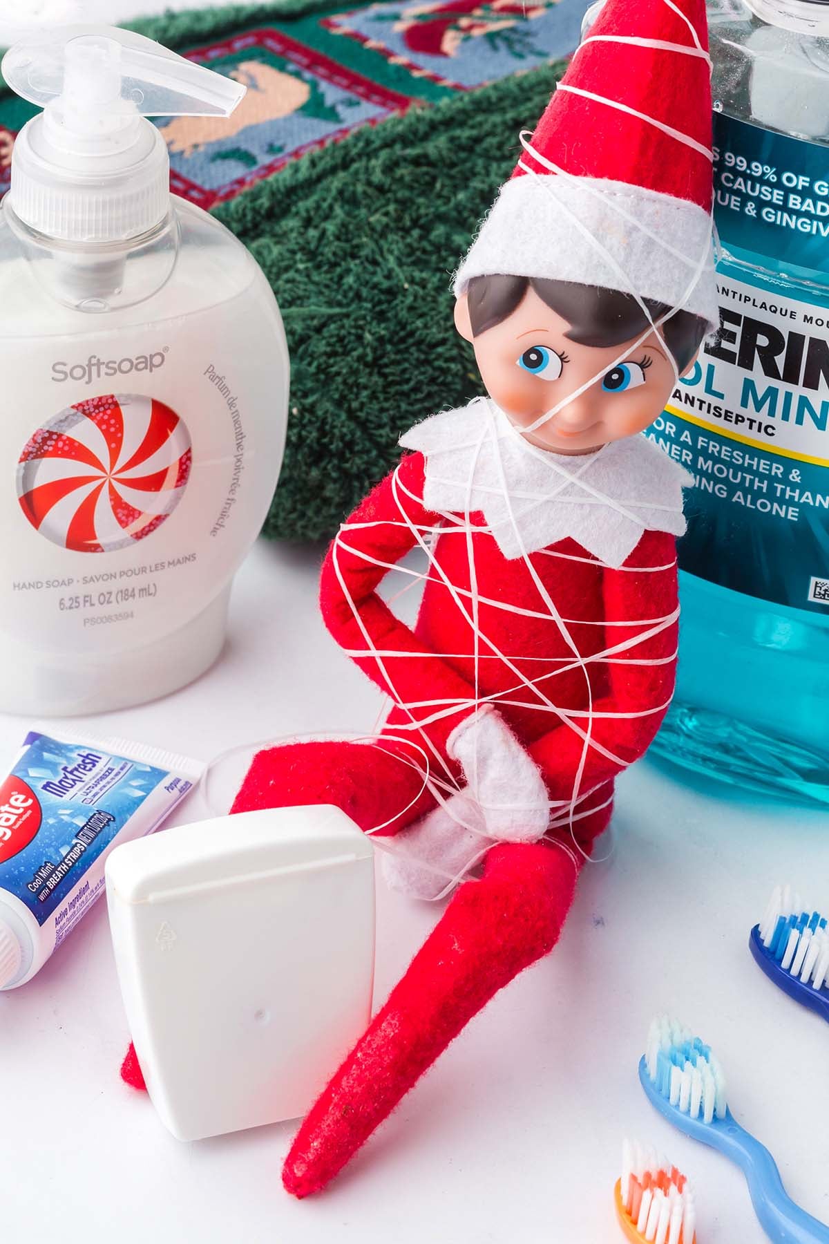 An elf doll wrapped in white dental floss, sitting among a bottle of hand soap, mouthwash, and toothbrushes.