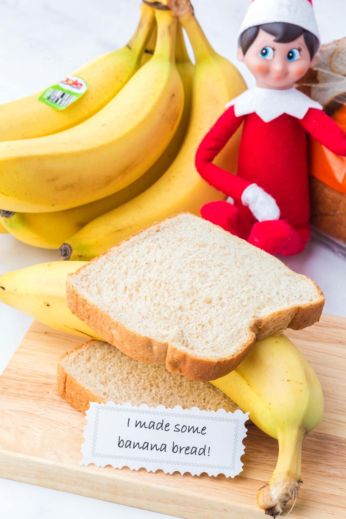 An Elf on the Shelf doll sits next to bananas and bread slices with a note reading “I made some banana bread!”.