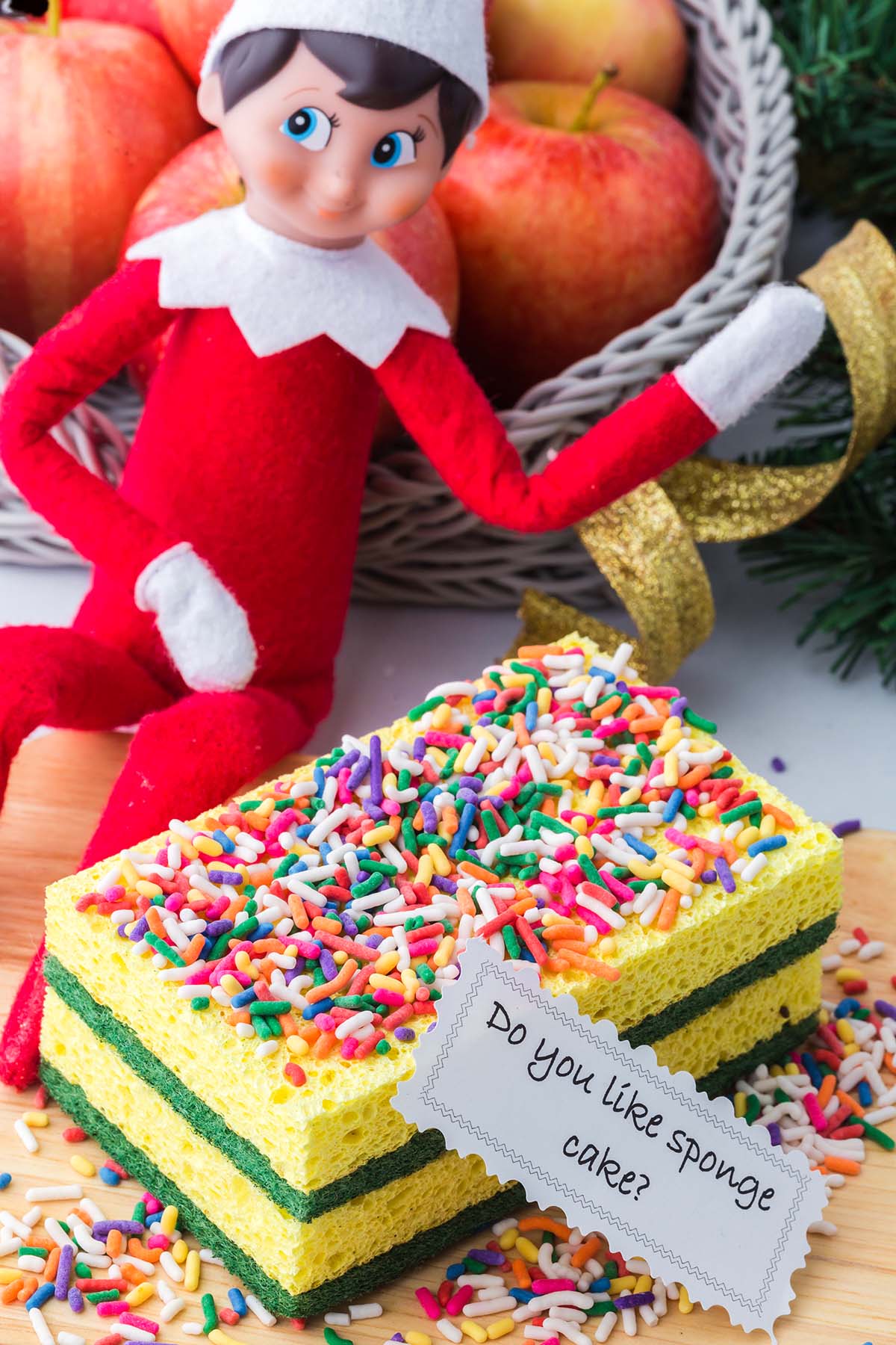An elf toy sits next to two stacked kitchen sponges decorated like cake with sprinkles and a note reading, “Do you like sponge cake?”.
