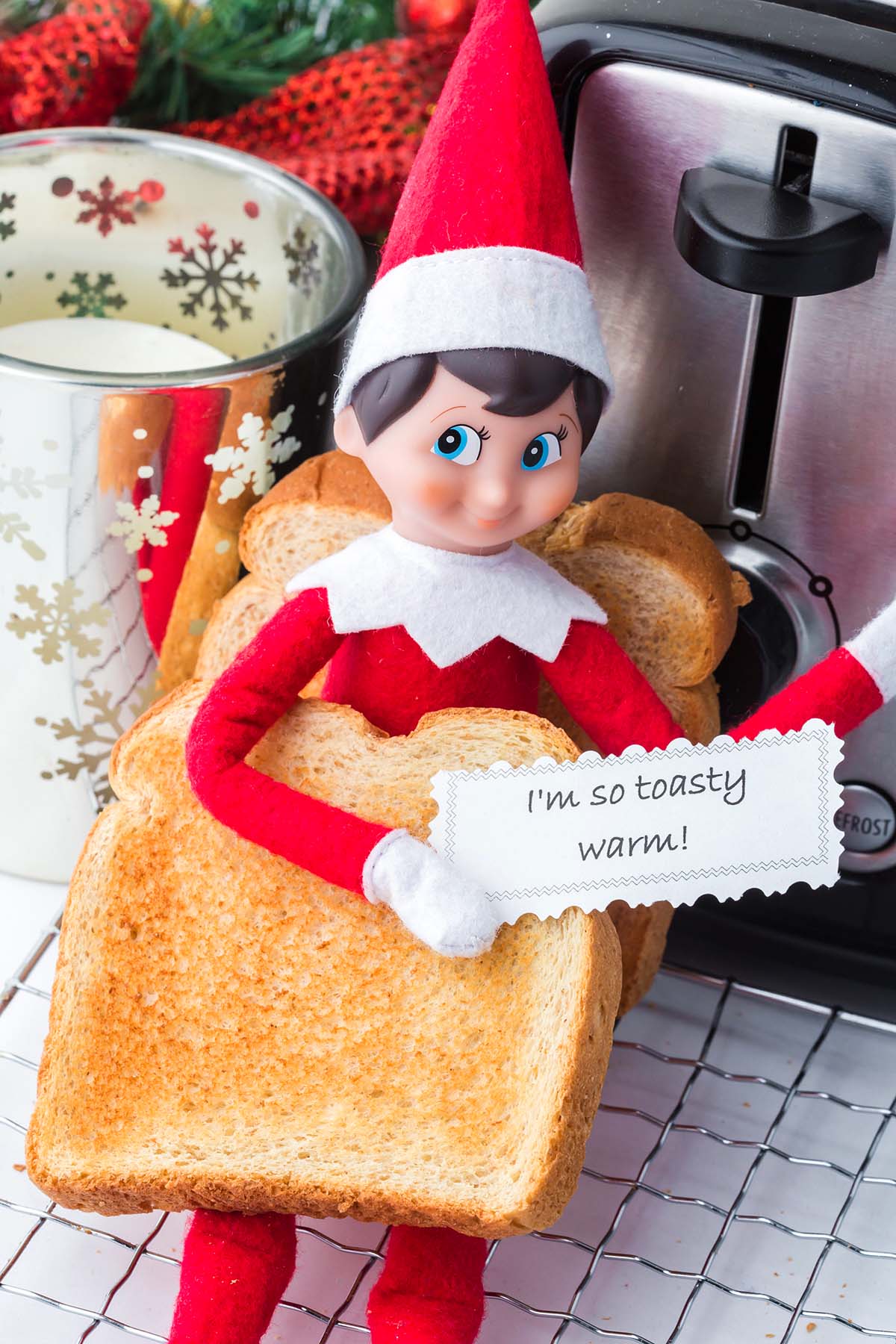 An Elf on the Shelf figurine holds a piece of toast with a note that reads, “I’m so toasty warm!”.