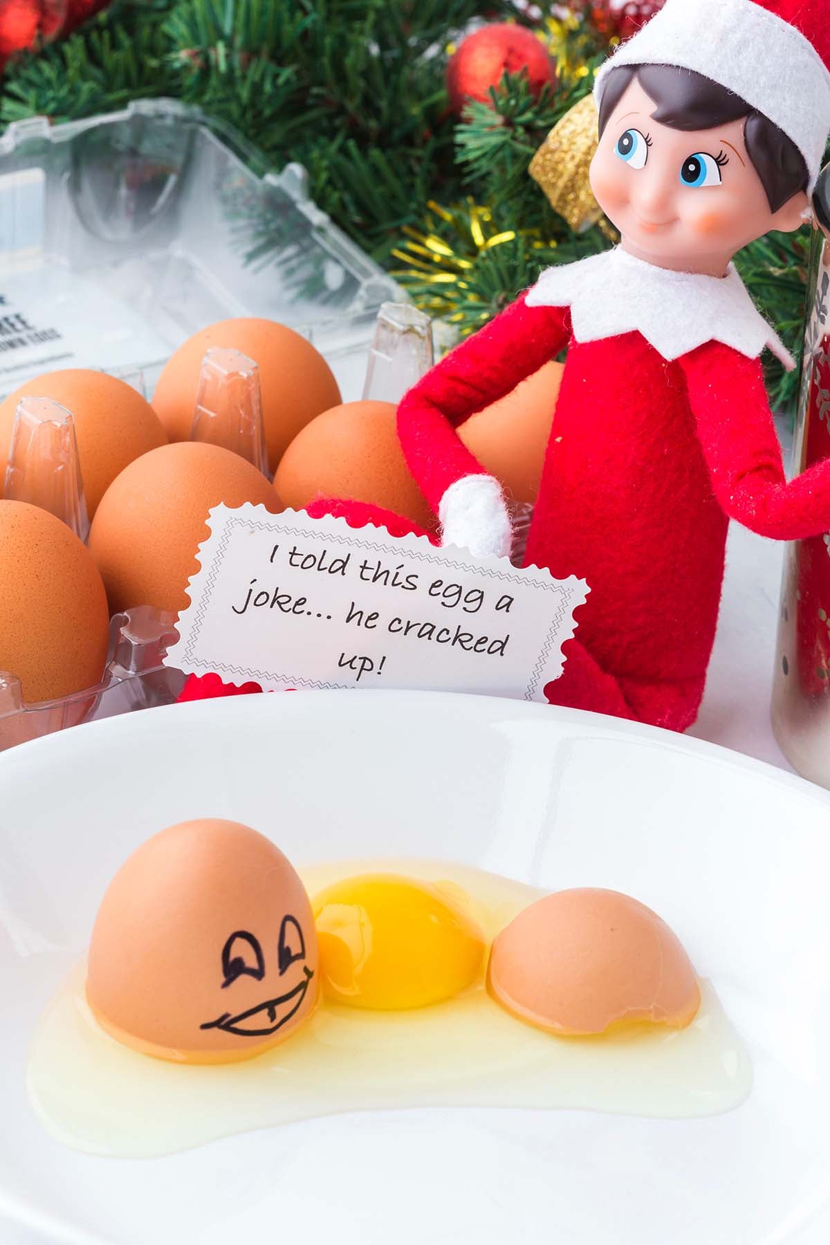 An Elf on the Shelf holding a note about a cracked egg, next to a bowl containing a cracked egg with a drawn-on face.