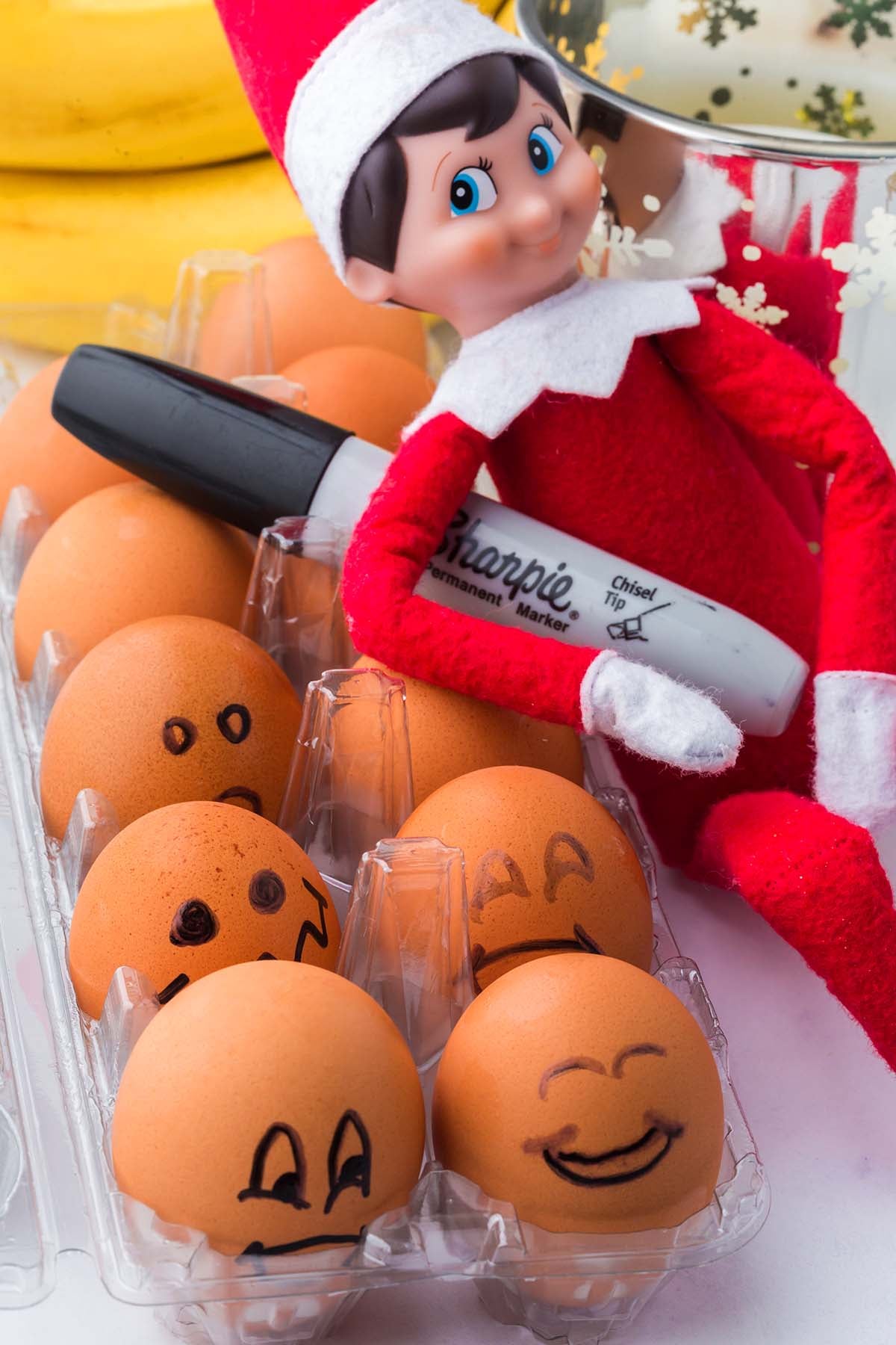 An Elf on the Shelf holding a black marker in a carton of brown eggs with drawn-on faces.