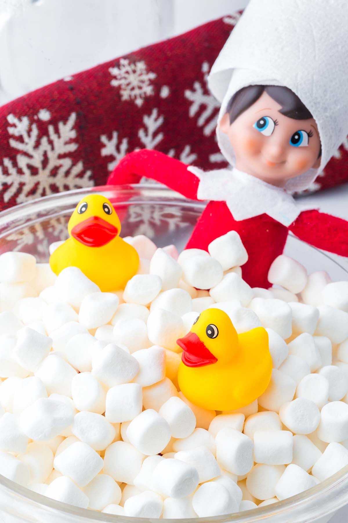 An elf doll and two yellow rubber ducks inside a glass bowl filled with marshmallows.