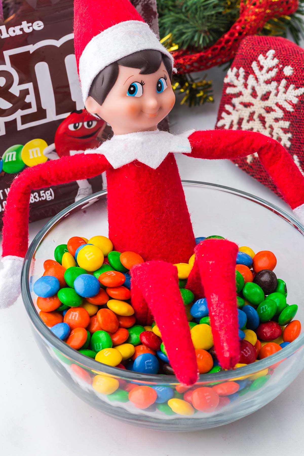 An Elf on the Shelf doll sitting inside a glass bowl filled with colorful M&M’s.