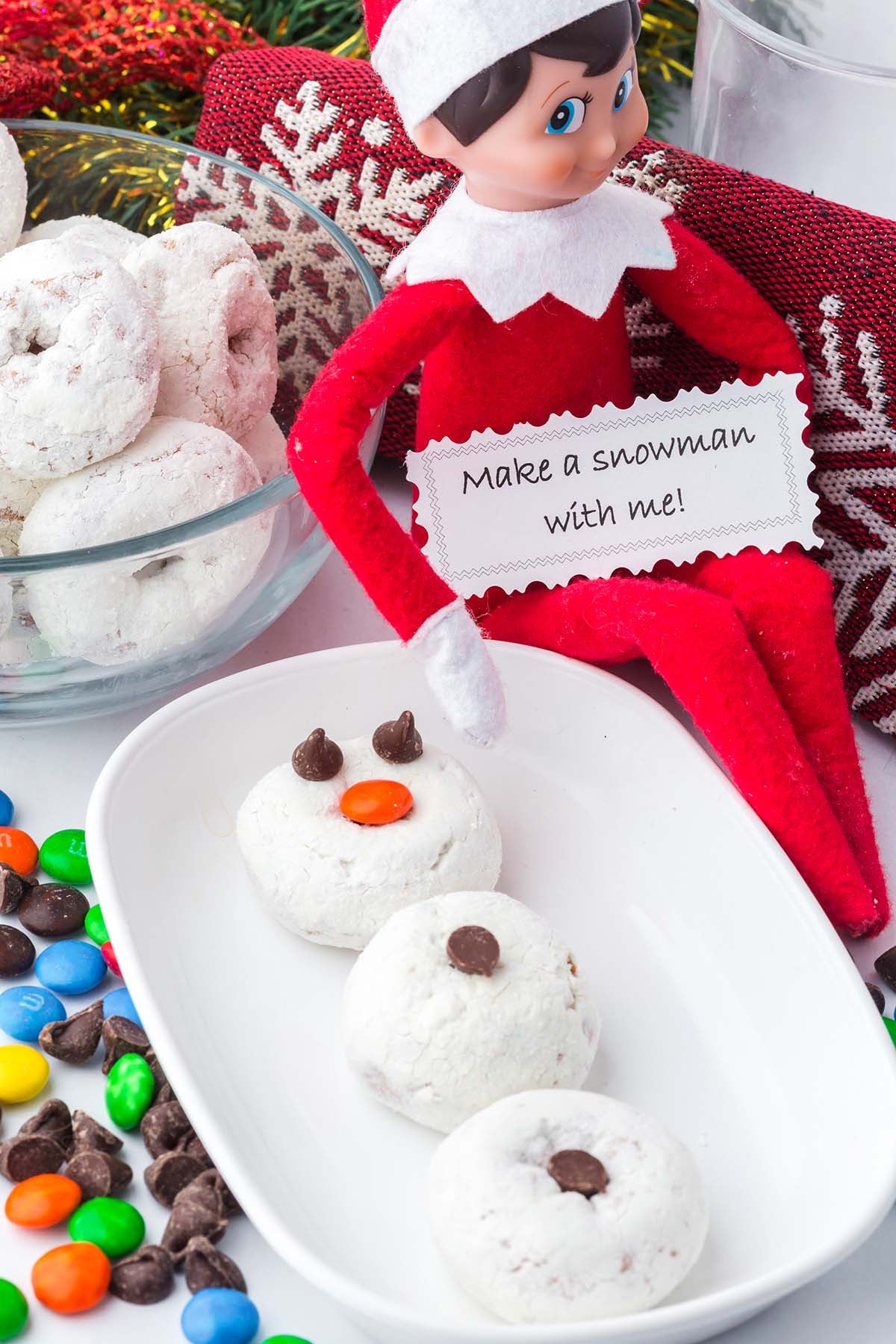 An Elf on the Shelf doll holds a note that says “Make a snowman with me!” next to powdered donut snowmen.