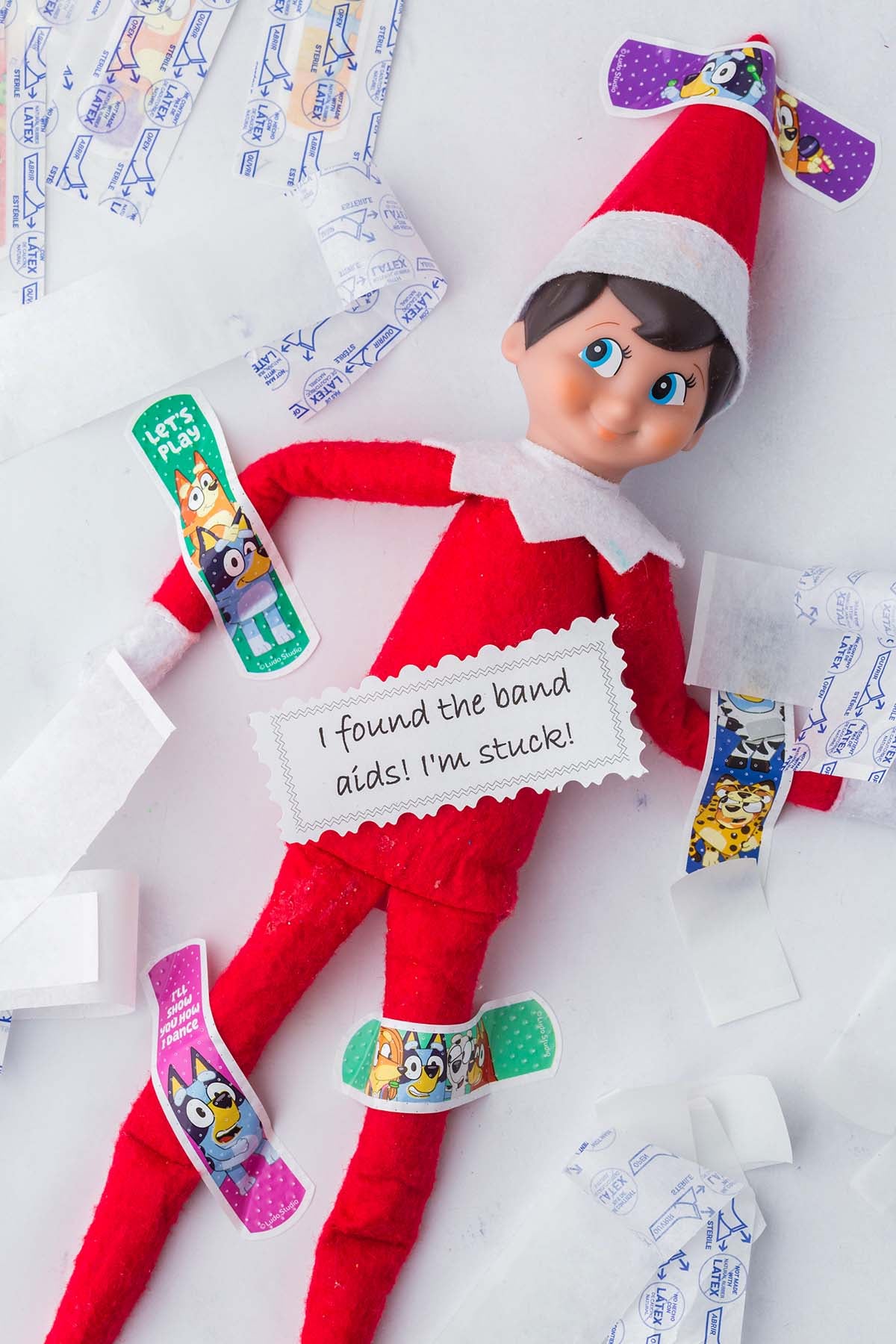 An Elf on the Shelf toy covered in Bluey-themed bandages with a note that reads, “I found the band aids! I’m stuck!”.