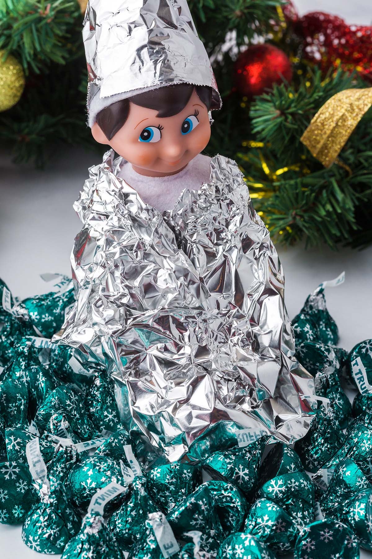 An elf doll wearing a foil hat and cape, sitting in a pile of green-wrapped chocolate candies.