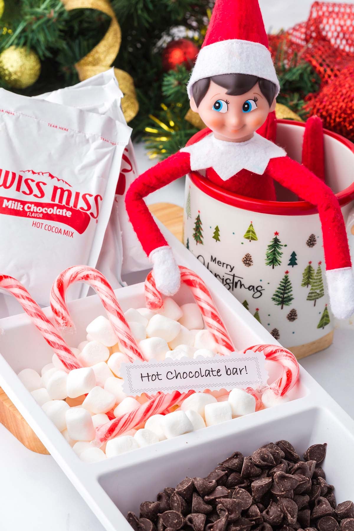 An Elf on the Shelf sits in a mug next to a tray of marshmallows, candy canes, and chocolate chips labeled Hot Chocolate.