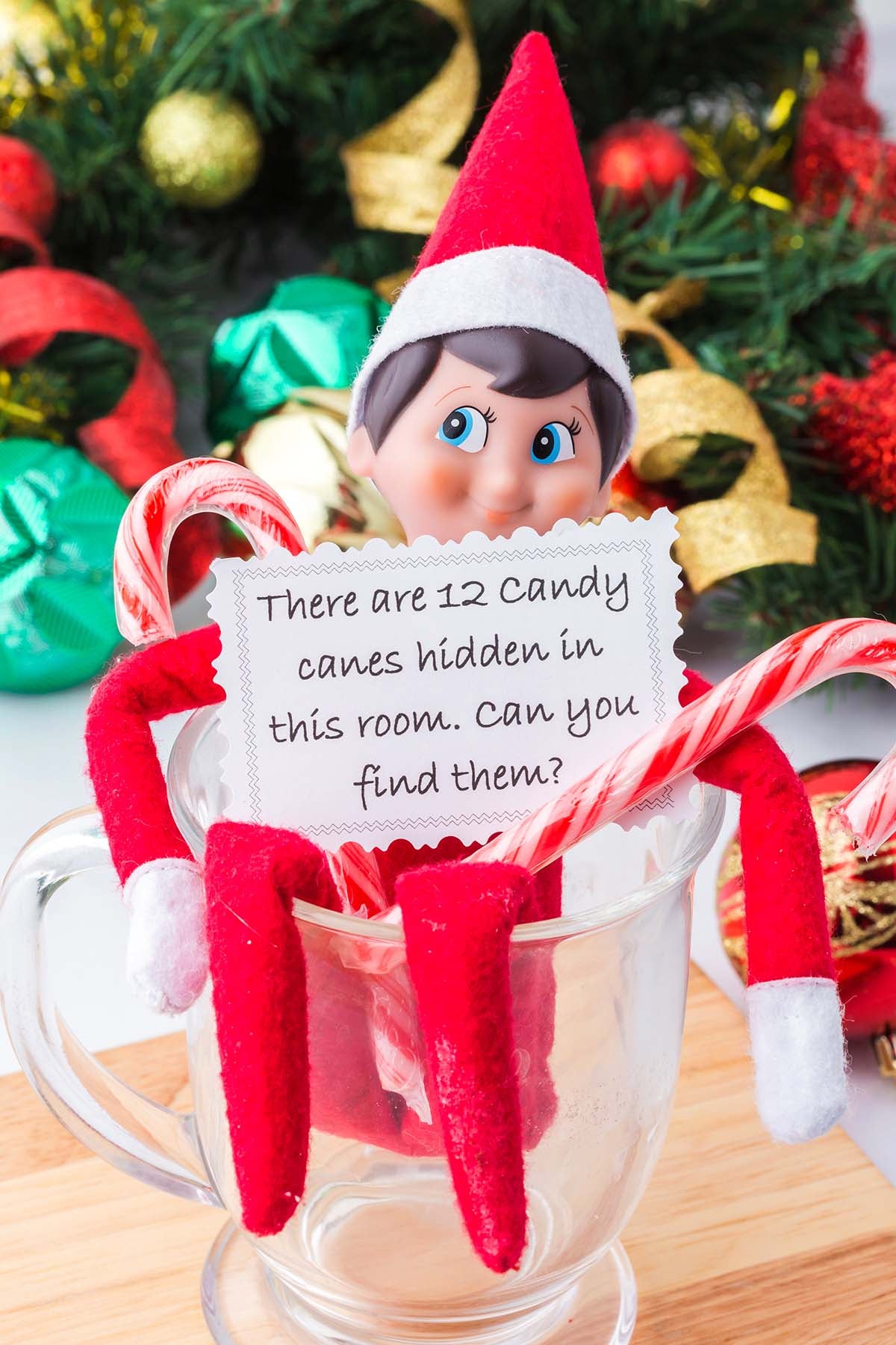 An elf doll in a glass mug holding a note that reads, “There are 12 candy canes hidden in this room. Can you find them?”.