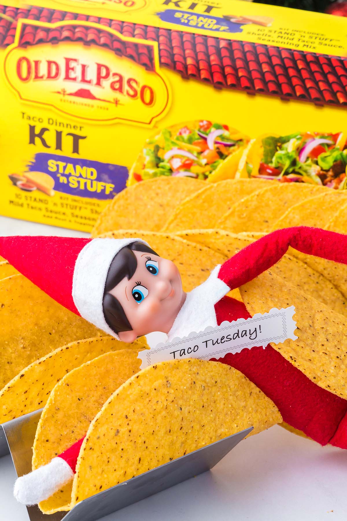 An Elf on the Shelf doll nestled among empty taco shells in front of an Old El Paso taco dinner kit.