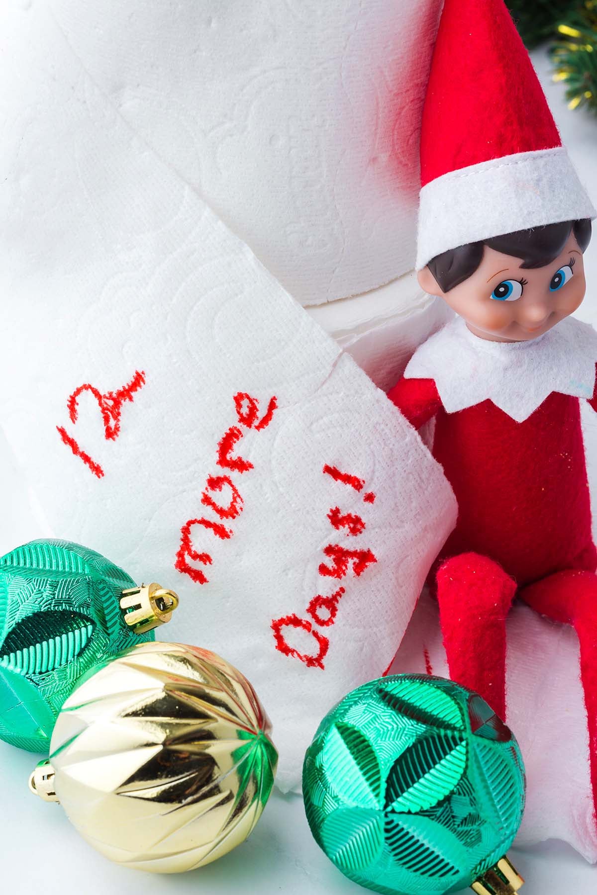 An elf doll sitting next to a roll of toilet paper with “12 more days!” written in red ink and three ornaments.