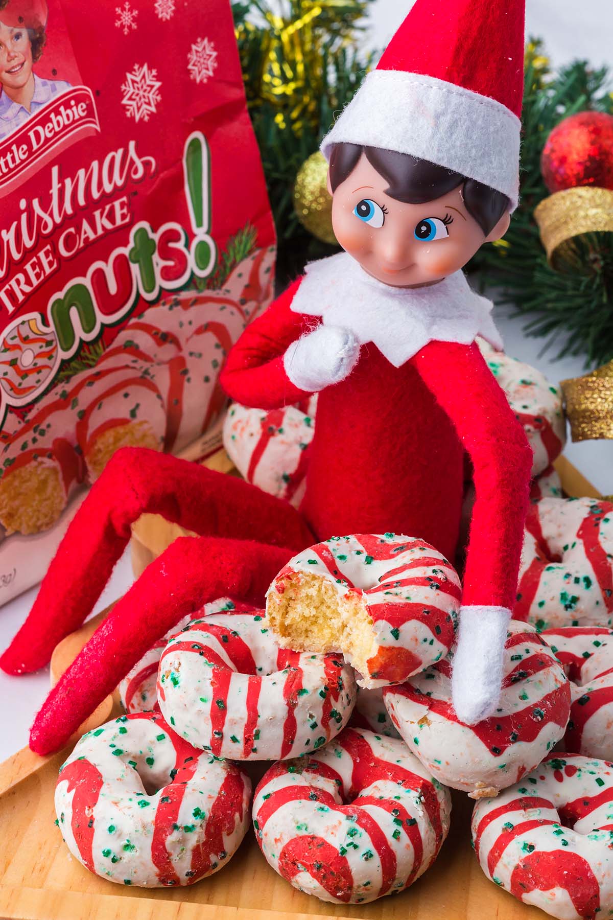 An Elf on the Shelf doll sits among a pile of Little Debbie Christmas Tree Cake donuts, holding one with a bite taken.