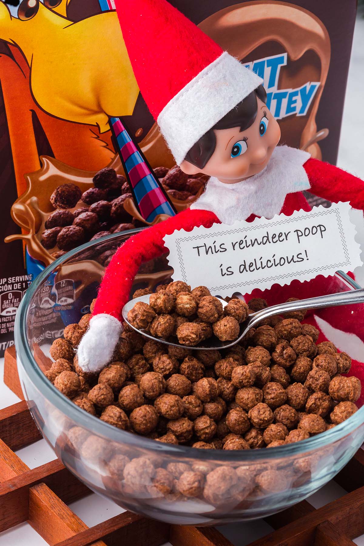 A toy elf holds a spoon over a bowl of chocolate cereal with a sign that reads, “This reindeer poop is delicious!”.