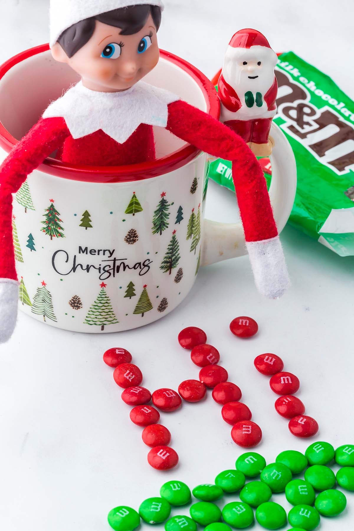 An elf doll sits in a “Merry Christmas” mug, with red M&Ms arranged to spell “Hi” on the table.