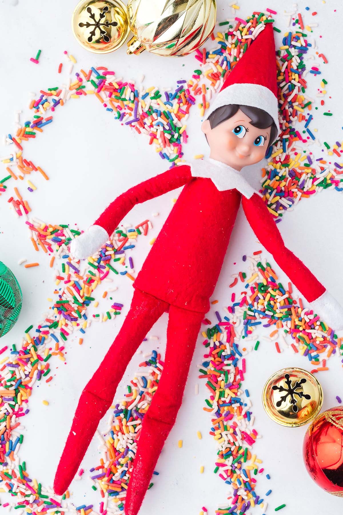 An Elf on the Shelf doll lying in a pile of colorful sprinkles with Christmas ornaments.