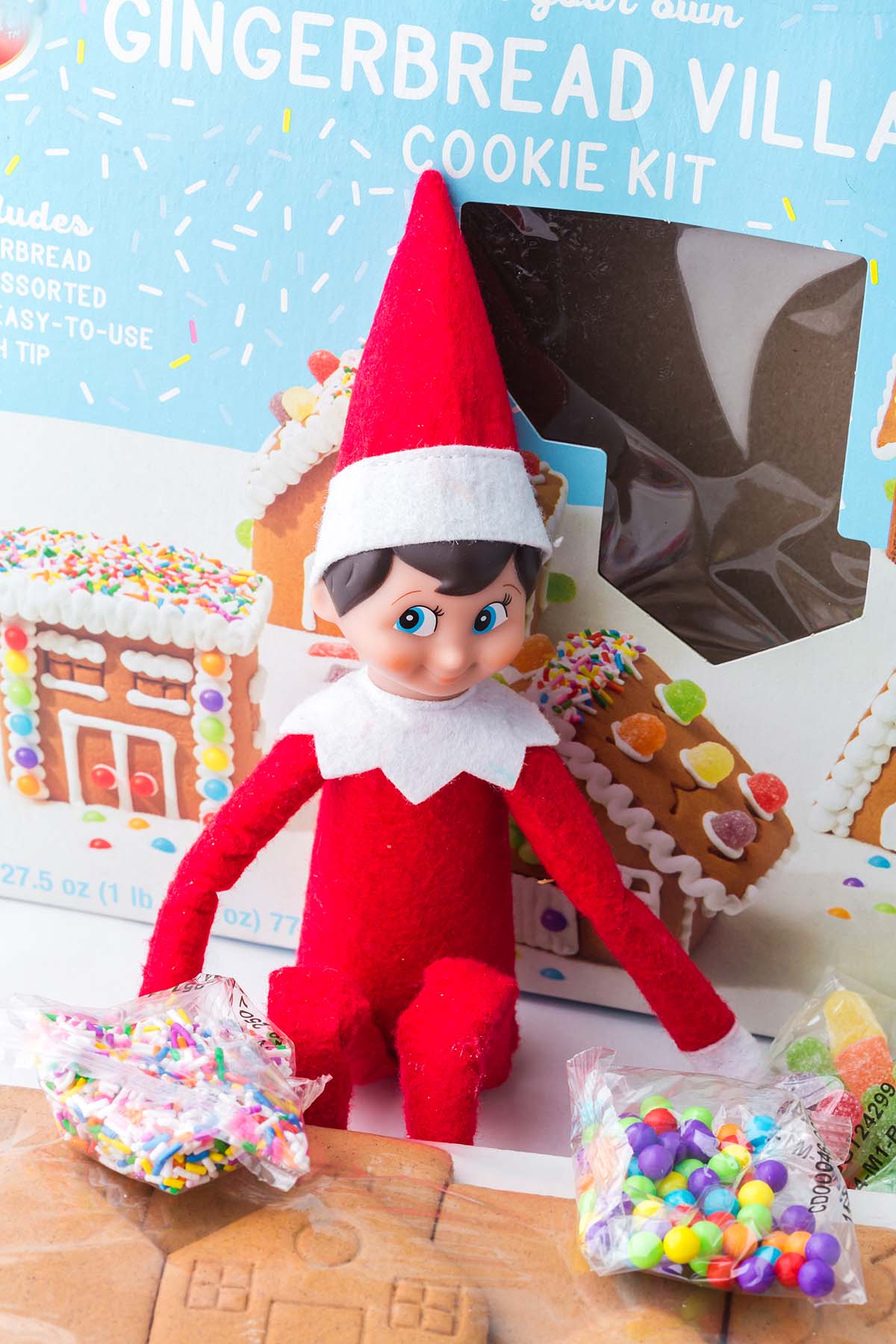 An Elf on the Shelf toy sitting in front of a gingerbread village cookie kit.