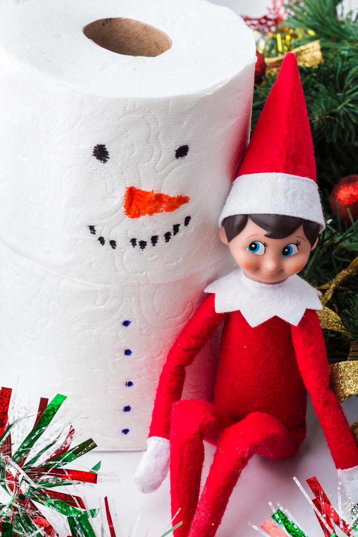An elf doll sitting next to a roll of toilet paper decorated to look like a snowman.
