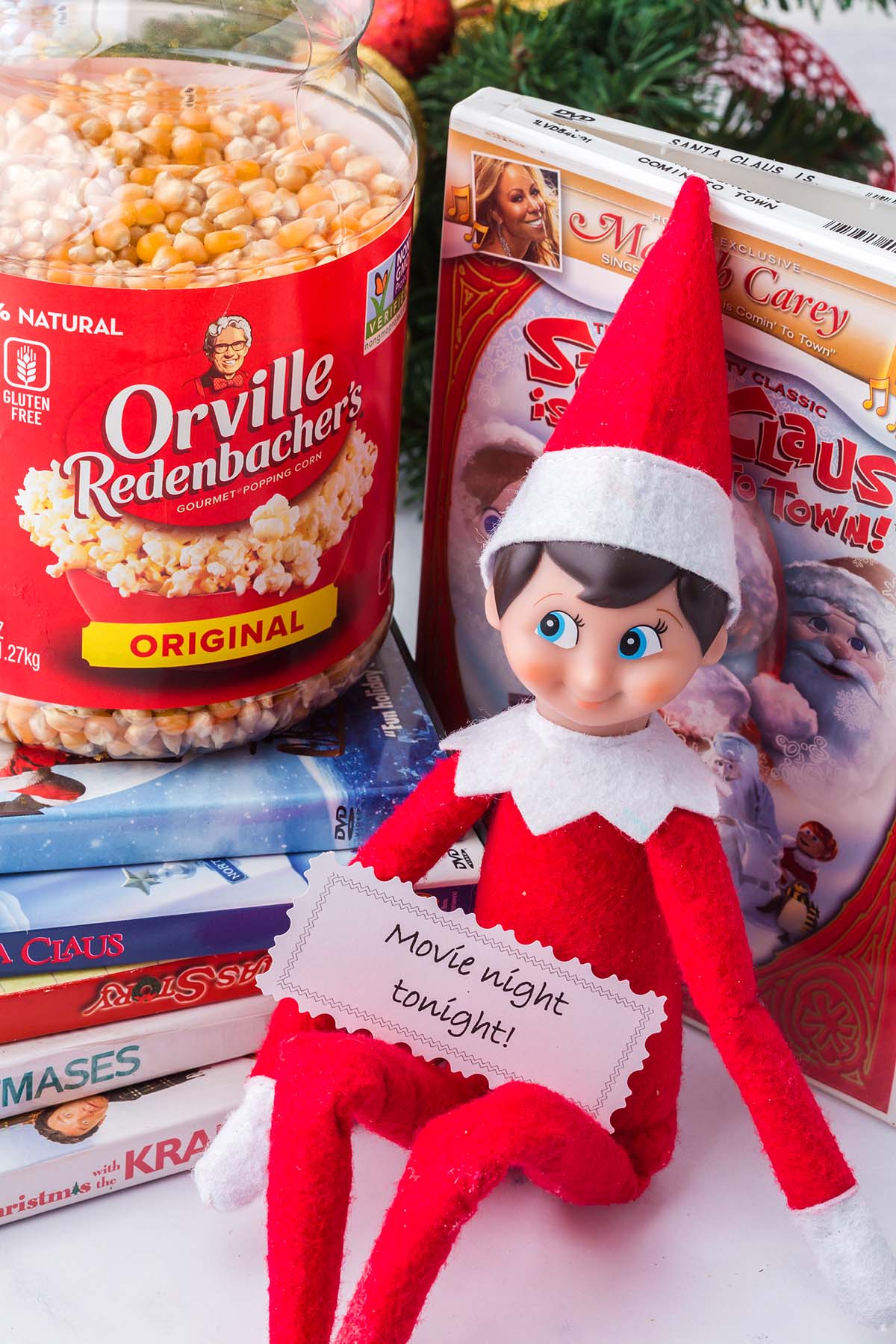 An Elf on the Shelf holding a note that says “Movie night tonight!” next to a jar of popcorn and Christmas DVDs.