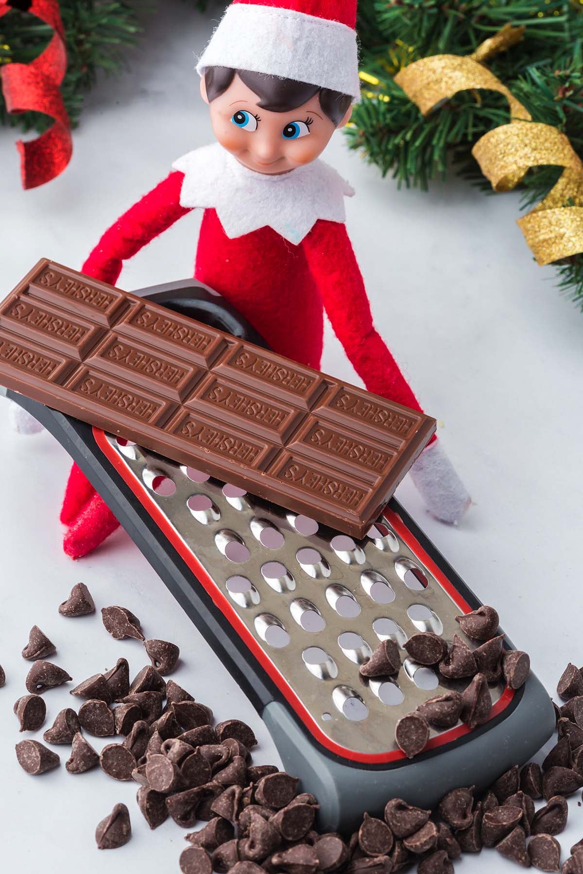 An Elf on the Shelf doll grating a Hershey’s chocolate bar over a pile of chocolate chips.