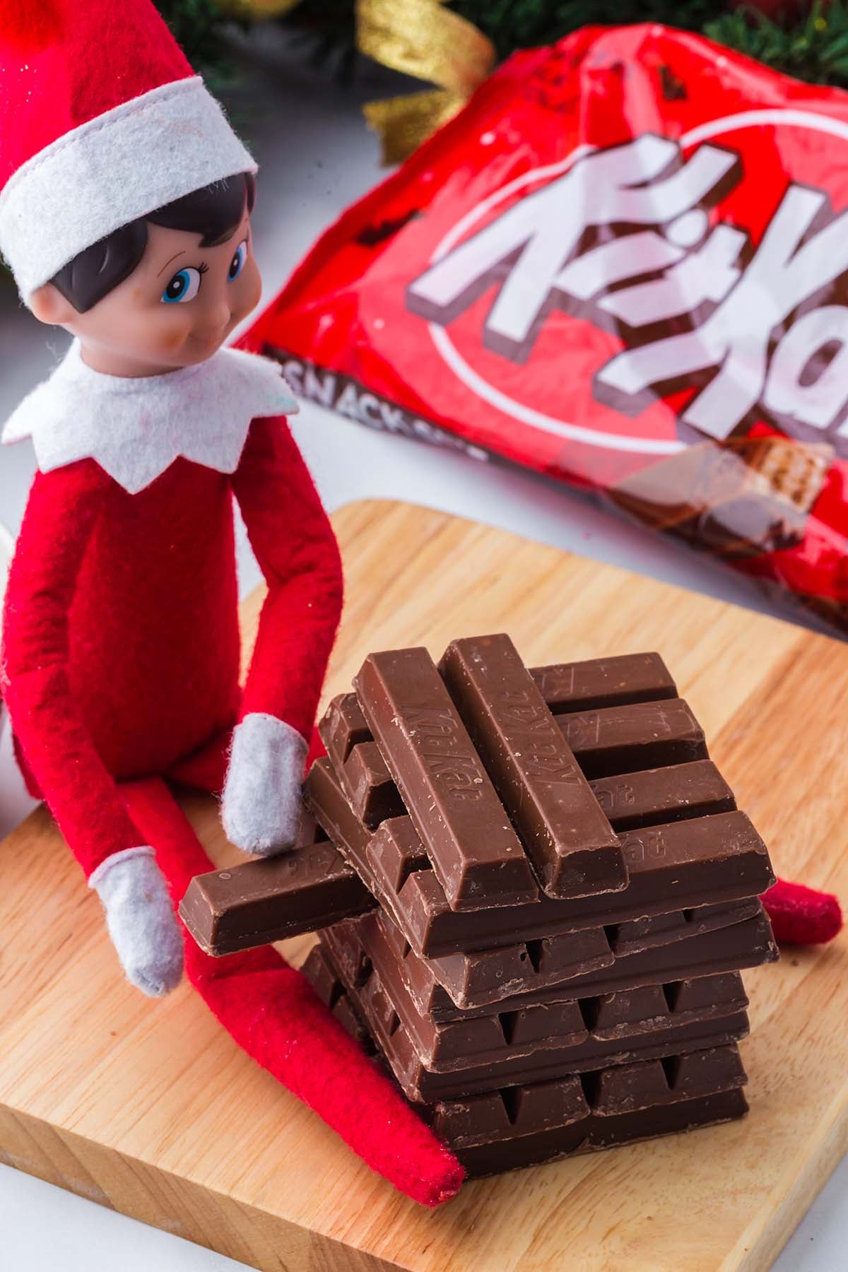 An Elf on the Shelf toy sitting next to a stack of Kit Kat bars on a wooden board.