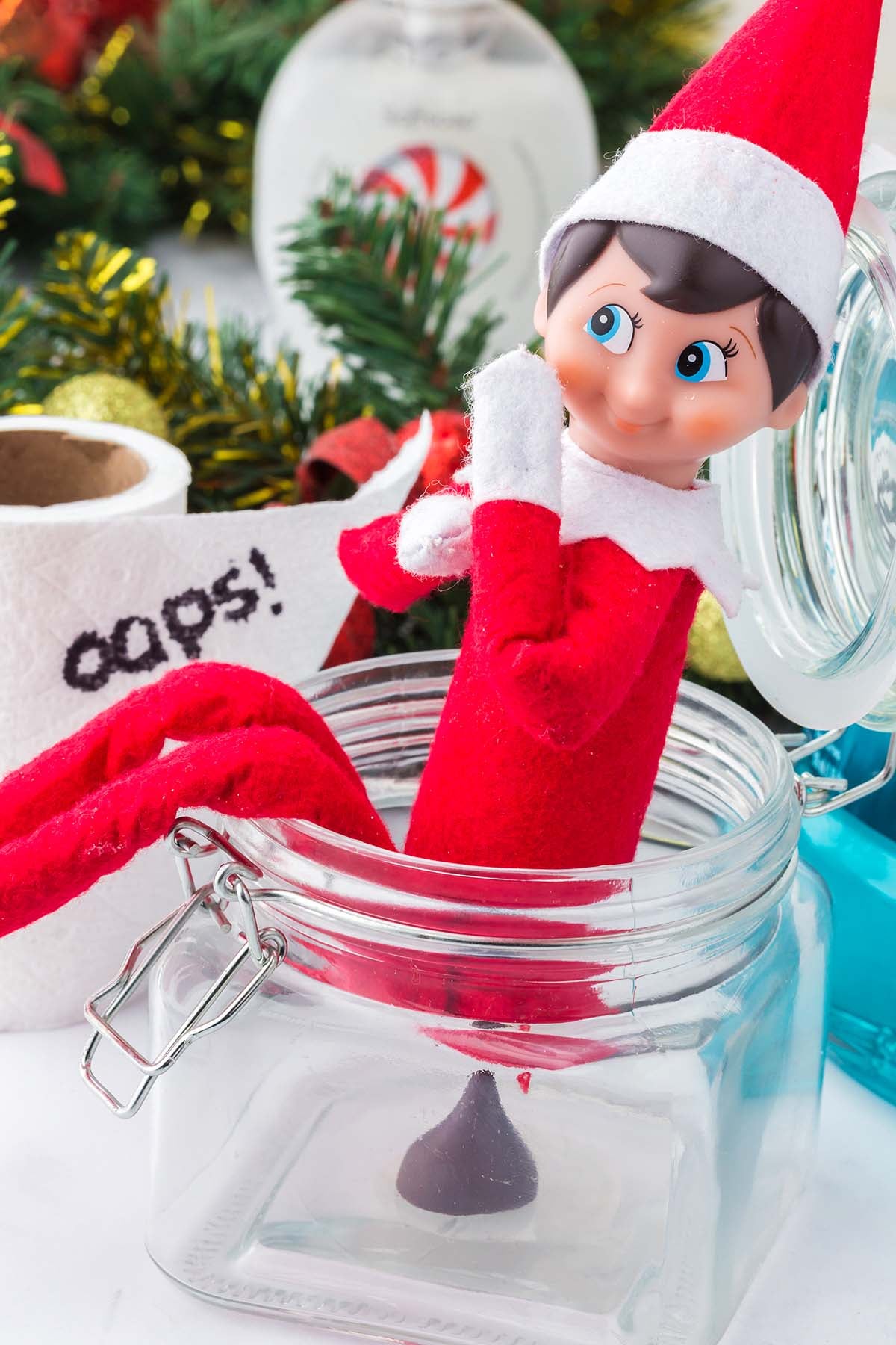 Elf on the Shelf sitting in a glass jar next to a roll of toilet paper with the word “oops!” written on it.