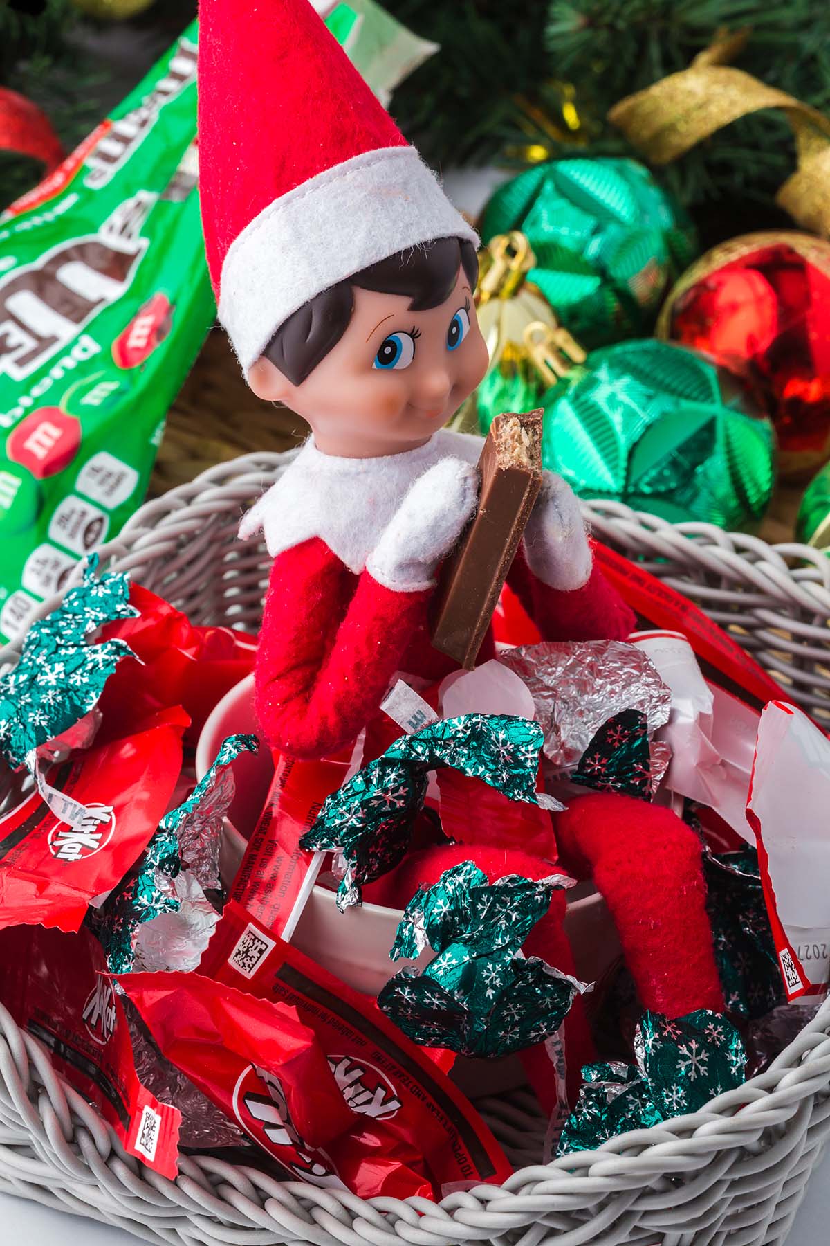An Elf on the Shelf doll sits in a basket filled with KitKat bars and candy wrappers, eating a piece of chocolate.