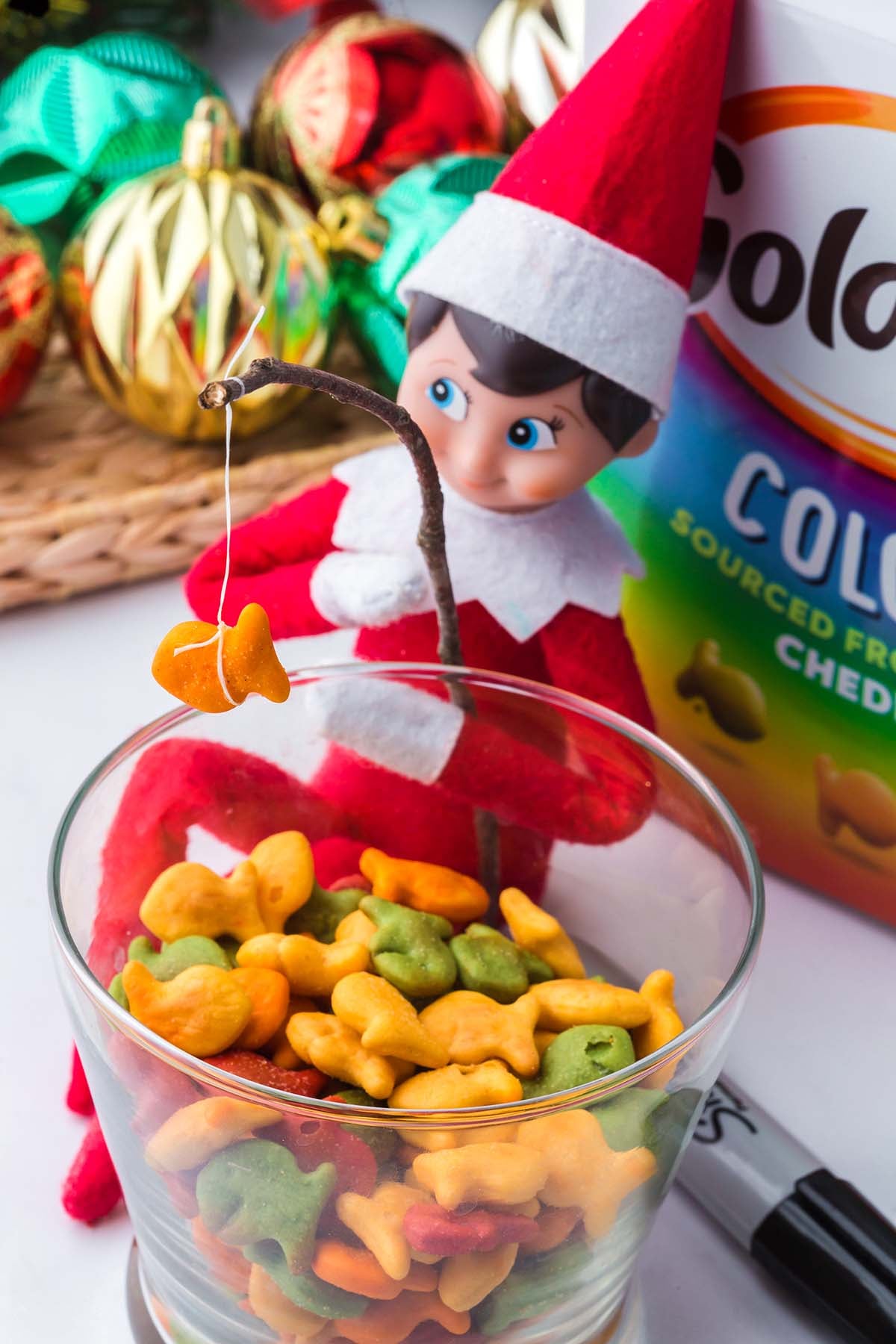 An elf doll sits on the rim of a glass bowl of Goldfish crackers, using a twig to fish a cracker out of the bowl.