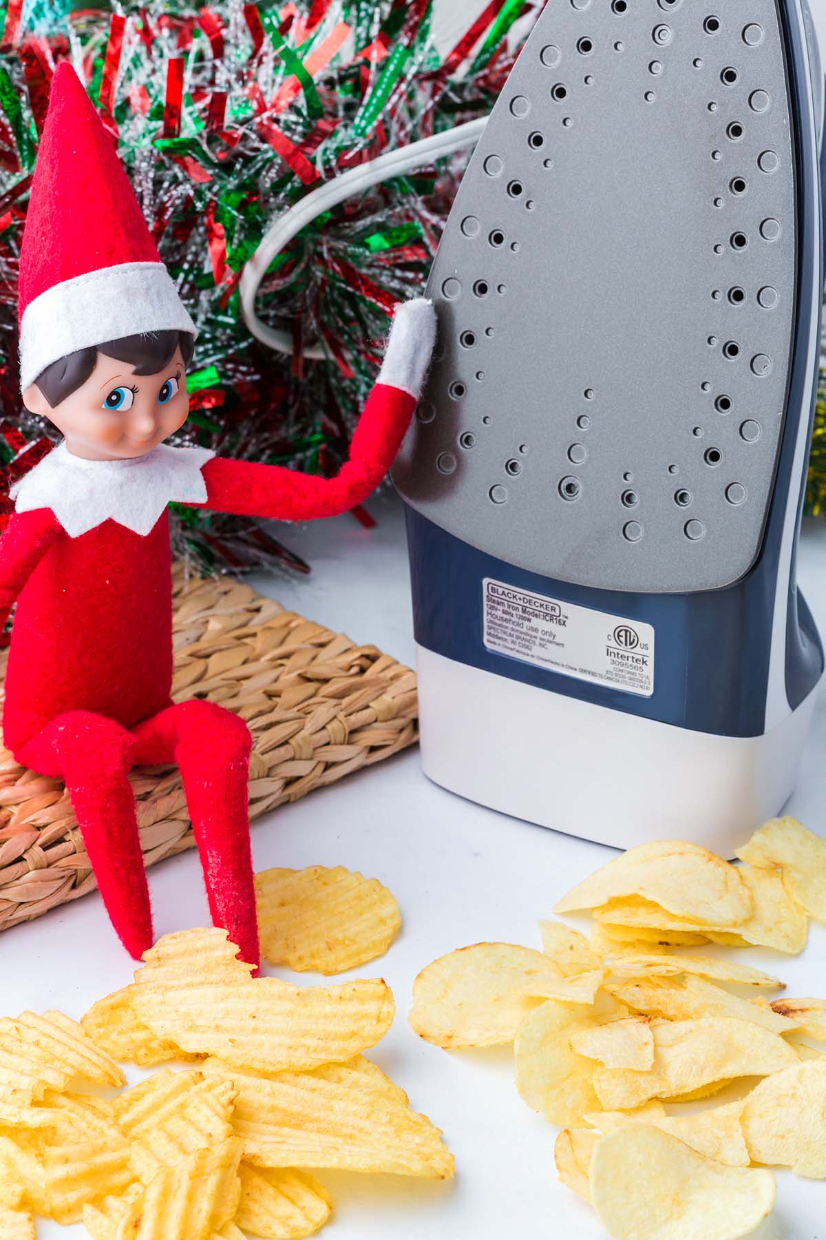 An Elf on the Shelf toy standing next to a clothing iron and a pile of potato chips.