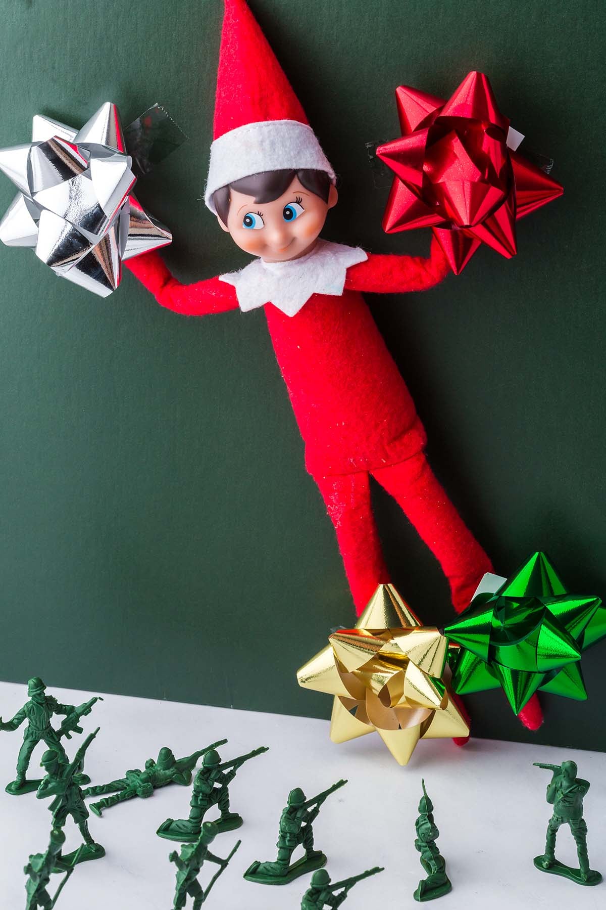 An Elf on the Shelf holding red and silver gift bows above a group of plastic green toy soldiers.