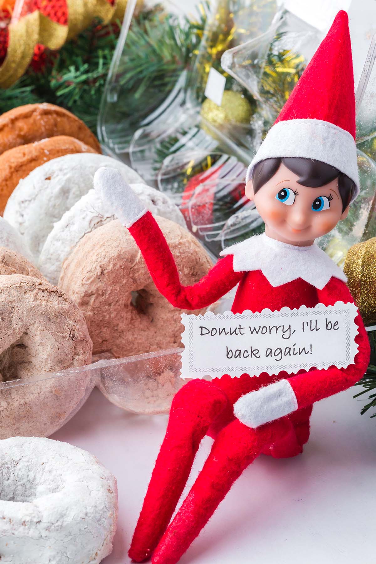 An elf toy holding a sign that says, “Donut worry, I’ll be back again!” next to a container of powdered donuts.