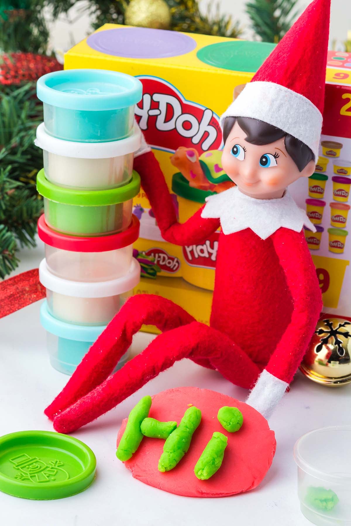 Elf on the Shelf figurine sitting next to a stack of Play-Doh cups and a red disc with “HI” spelled in green dough.