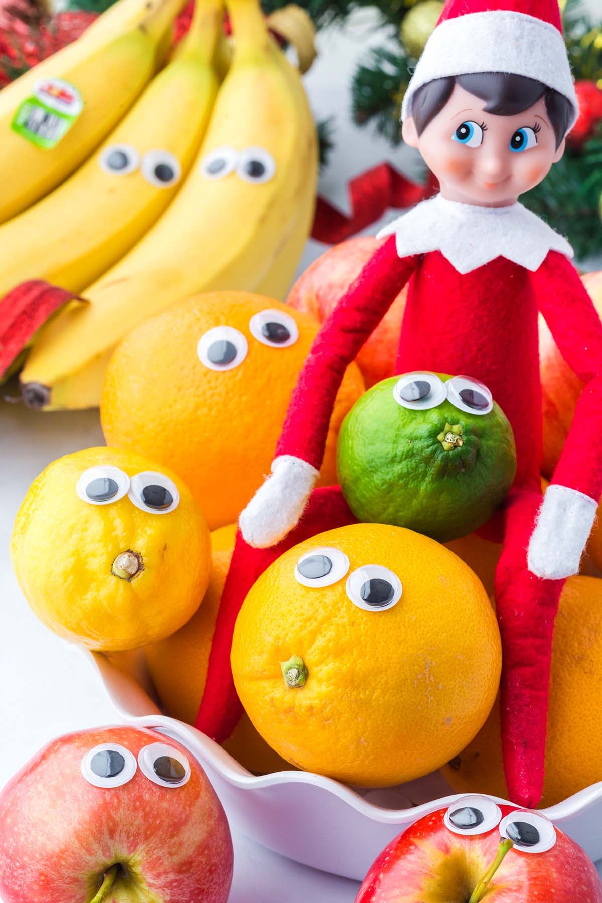 An elf doll sits in a bowl of fruit decorated with googly eyes.