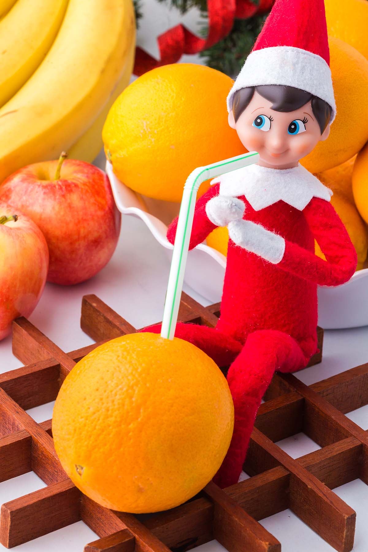 An Elf on the Shelf doll sits on a wooden trivet, drinking from an orange with a straw.